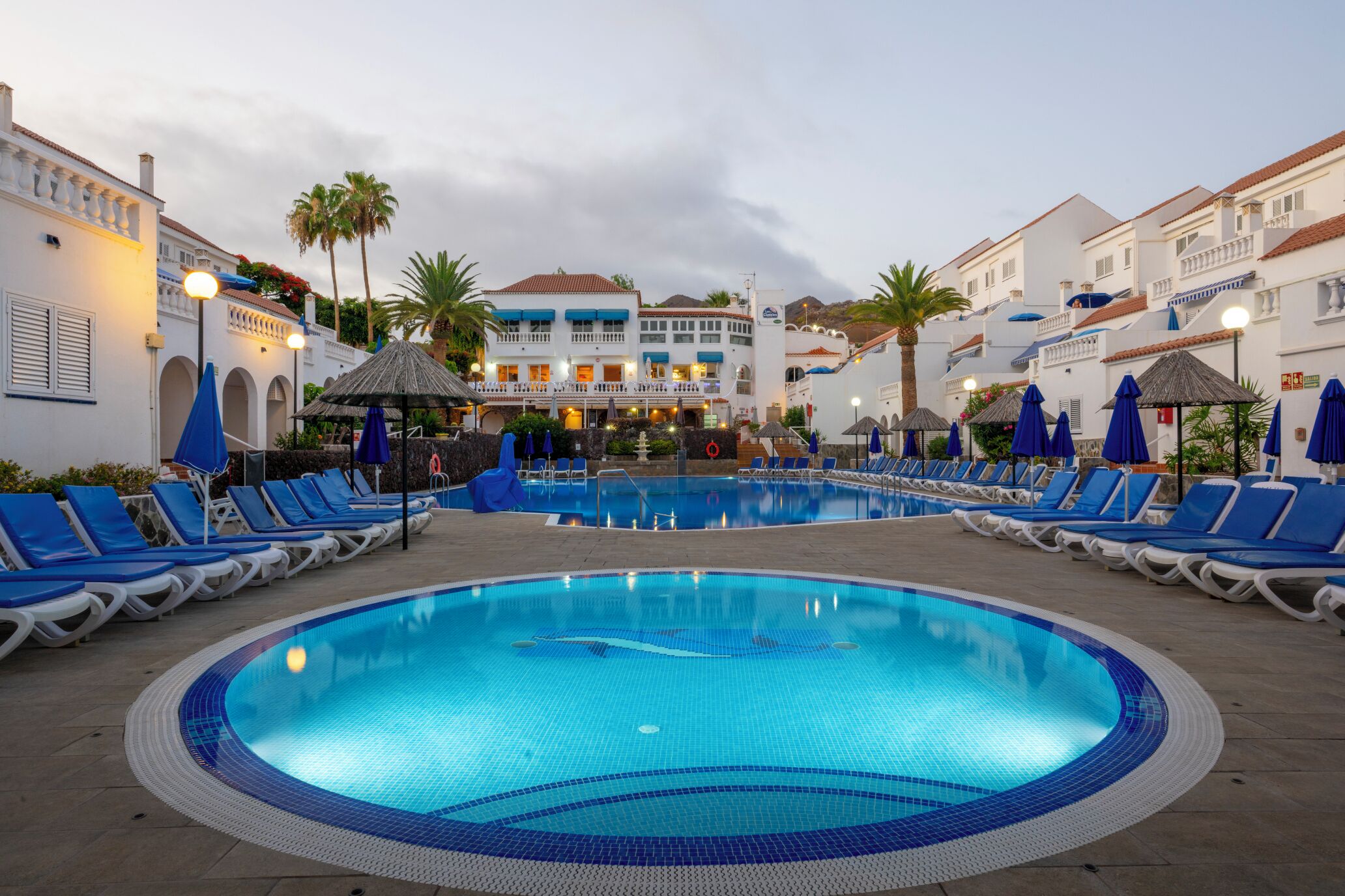 Une piscine entourée de bâtiments dotés de balcons, avec des chaises longues et des parasols à proximité.