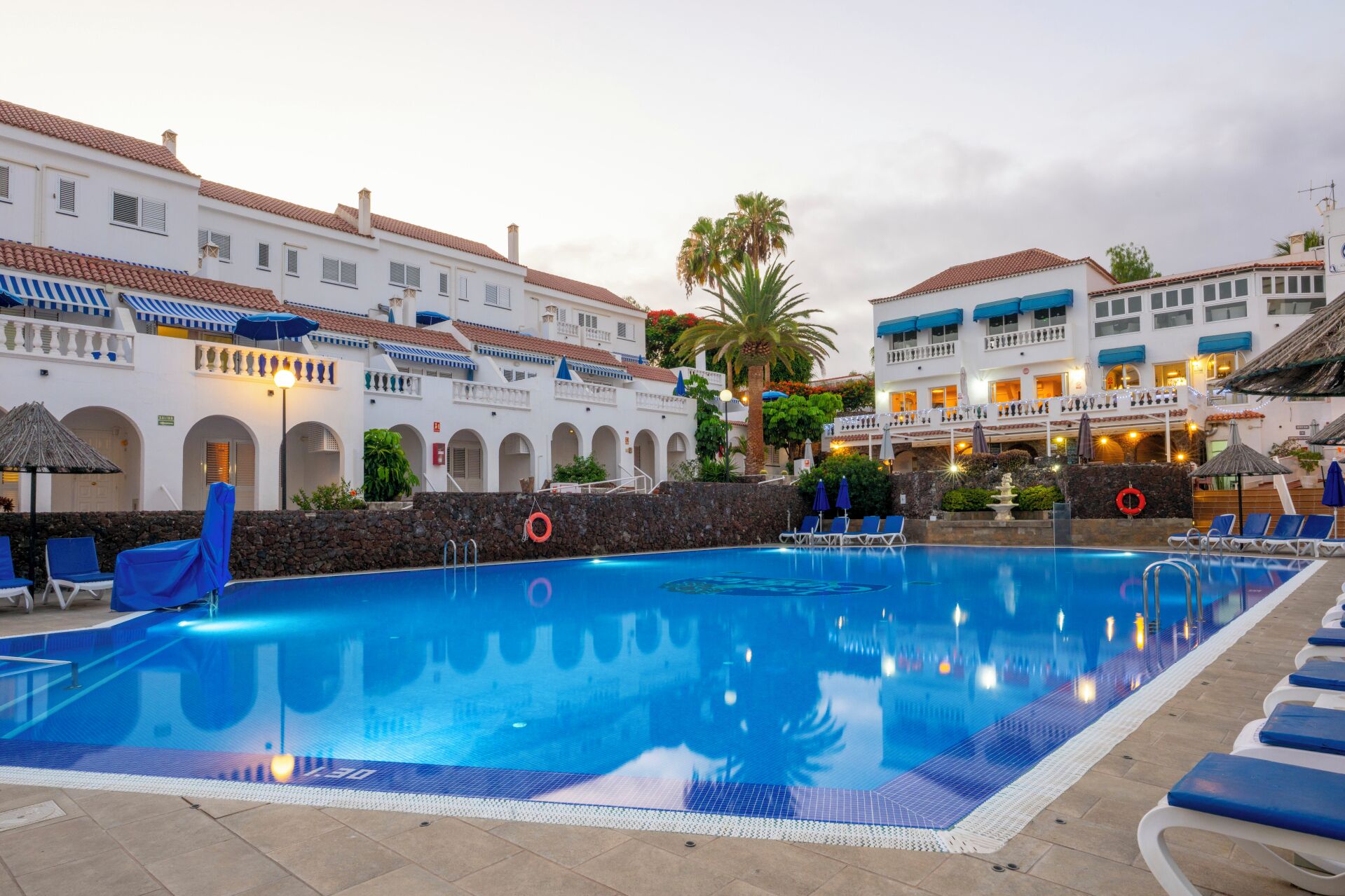 A swimming pool with lounge chairs and umbrellas, surrounded by white buildings with red-tiled roofs.