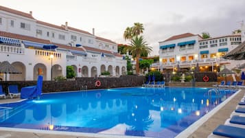 A swimming pool with lounge chairs and umbrellas, surrounded by white buildings with red-tiled roofs.