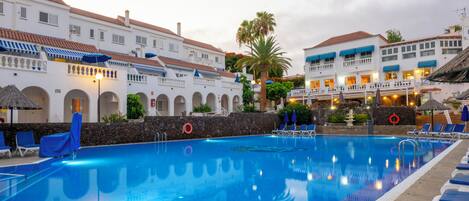A swimming pool with lounge chairs and umbrellas, surrounded by white buildings with red-tiled roofs.