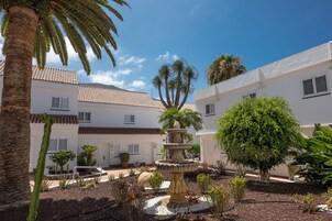 Un patio central con una fuente, rodeado de edificios blancos con techos de tejas rojas, palmeras y diversas plantas.