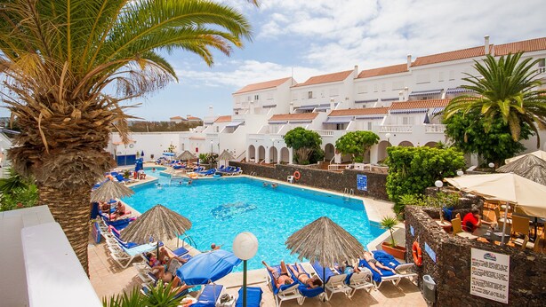 A pool area with lounge chairs and umbrellas, surrounded by palm trees and a building with red-tiled roofs. - Ona los Claveles (Arona)
