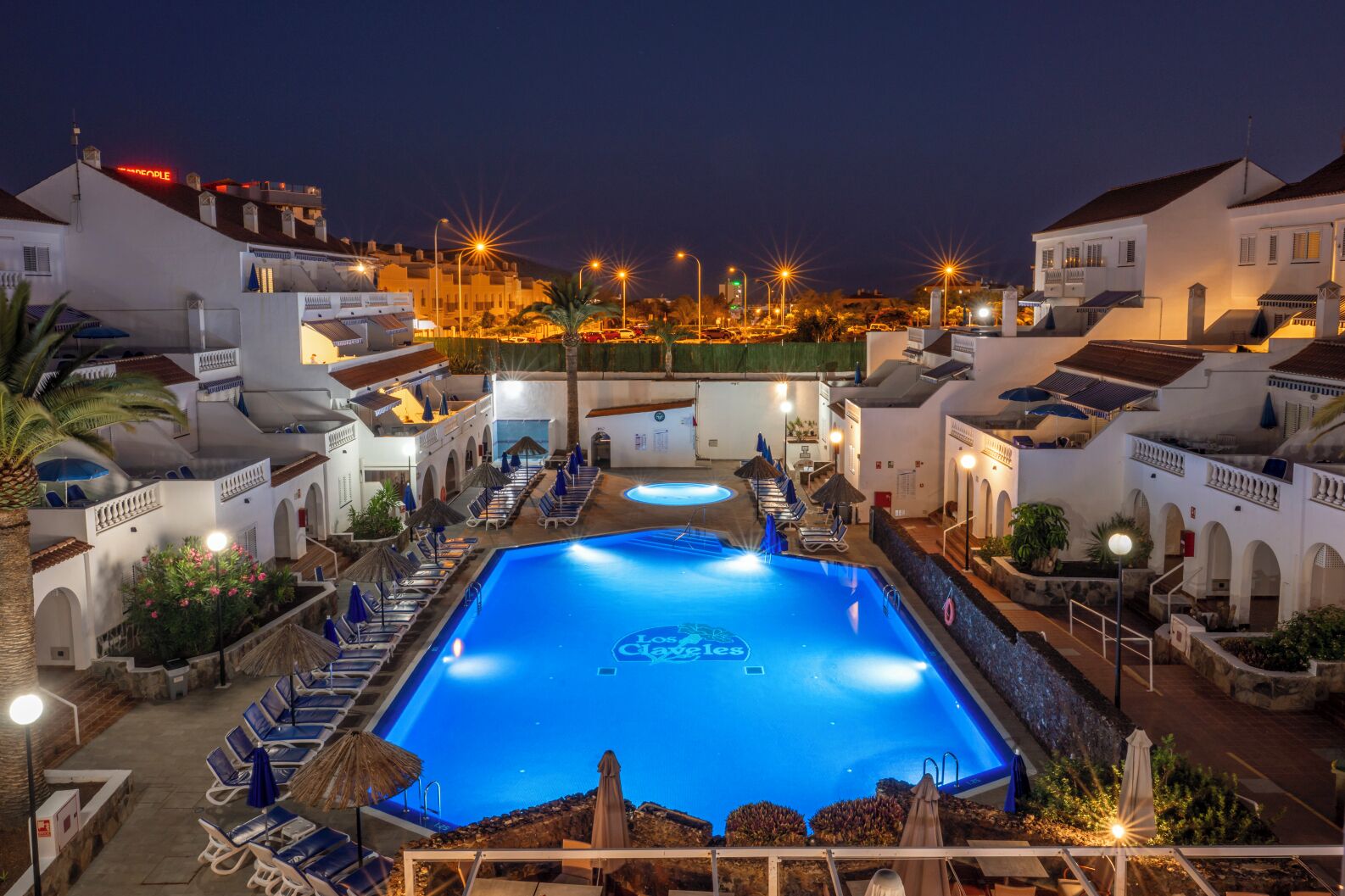 A hotel pool area with multiple lounge chairs and umbrellas, surrounded by white buildings with balconies.