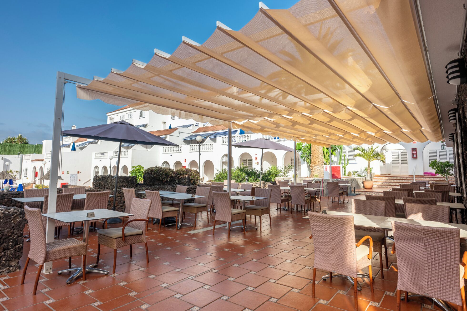 An outdoor café area with tables and chairs, shaded by large umbrellas, and a white building with arches in the background.