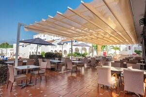 An outdoor café area with tables and chairs, shaded by large umbrellas, and a white building with arches in the background.