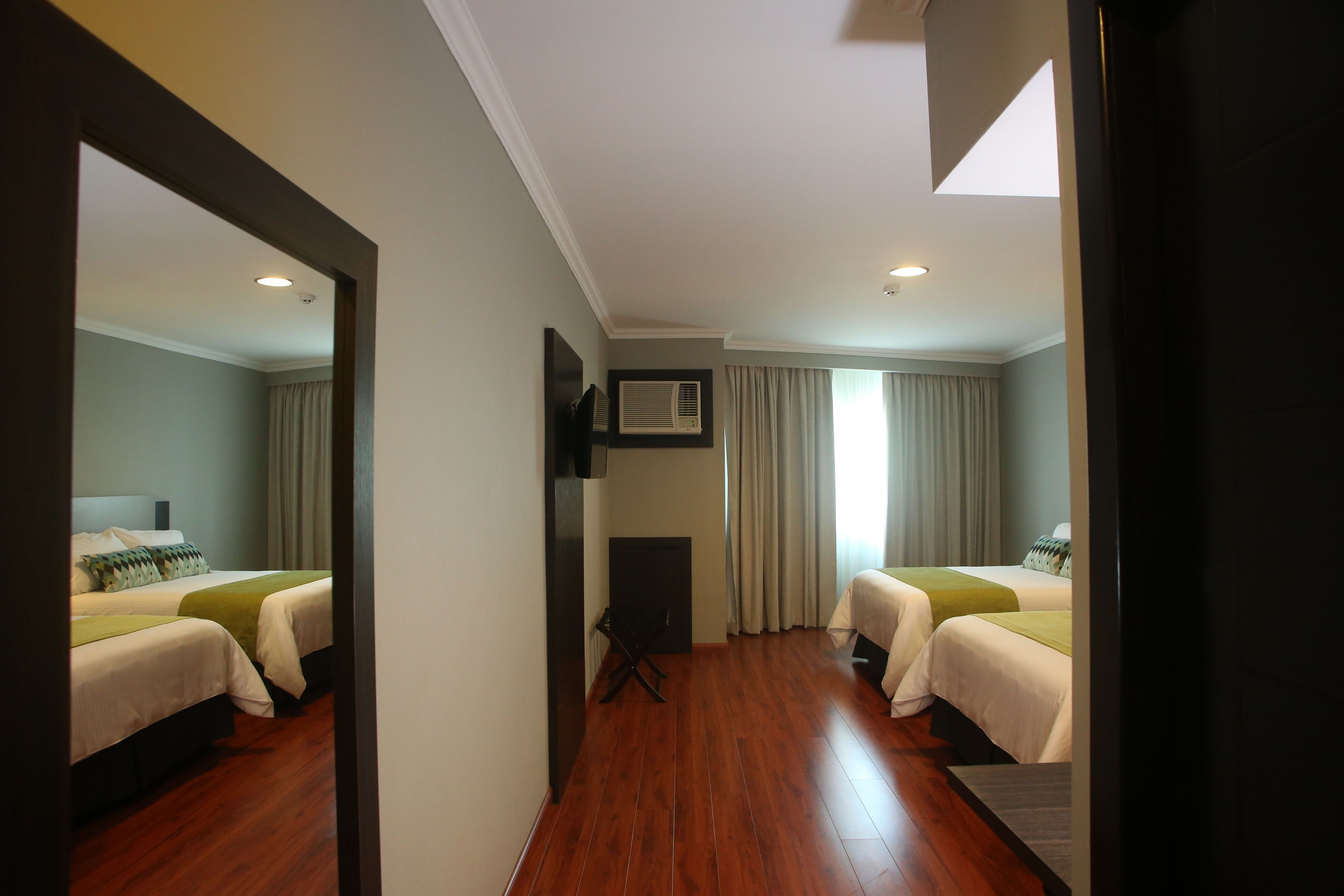 A hotel room with two beds, a flat-screen TV, an air conditioning unit, and a view of the outside through a window.