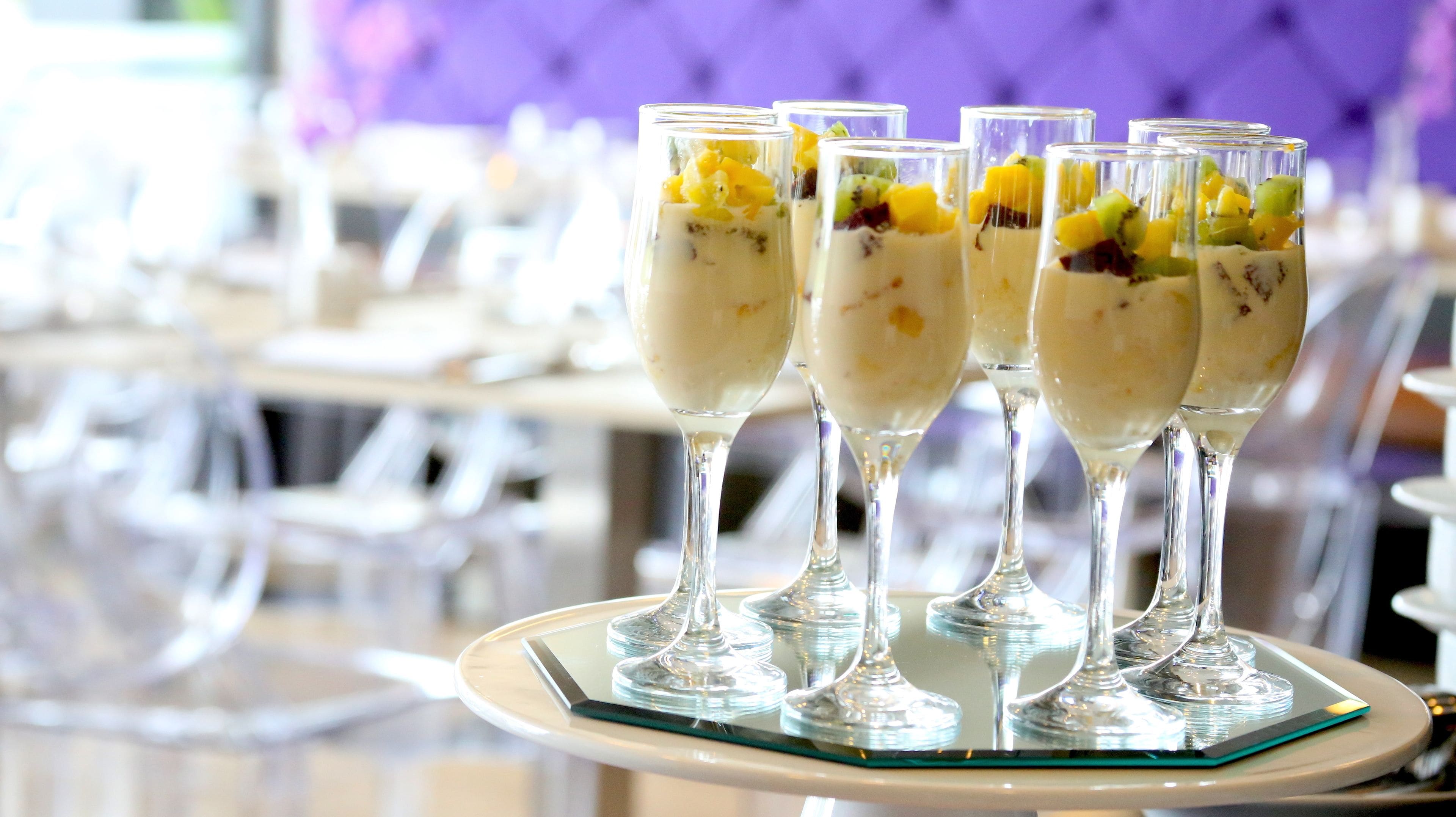 Four dessert glasses on a tray, likely in a dining area with white chairs and a purple wall in the background.