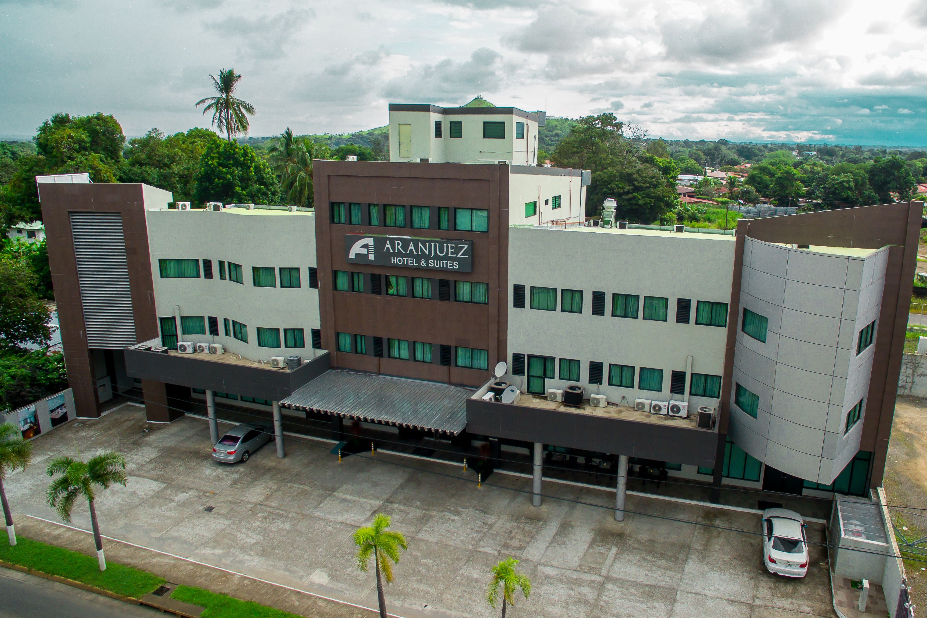 A hotel with a modern design, featuring a prominent sign for 'ARANJUEZ HOTEL & SUITES'. There are multiple parking spaces and a covered area for vehicles.