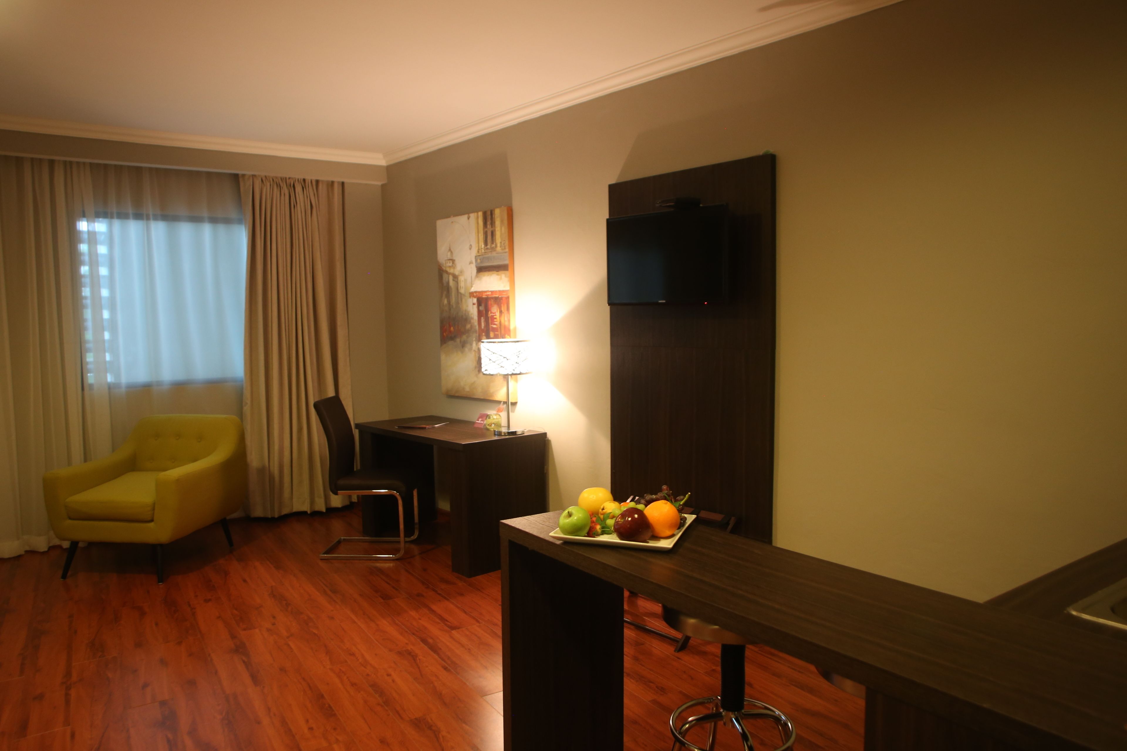 A hotel room with a wooden floor, a yellow armchair, a desk with a lamp, a flat-screen TV, and a fruit bowl on the desk.