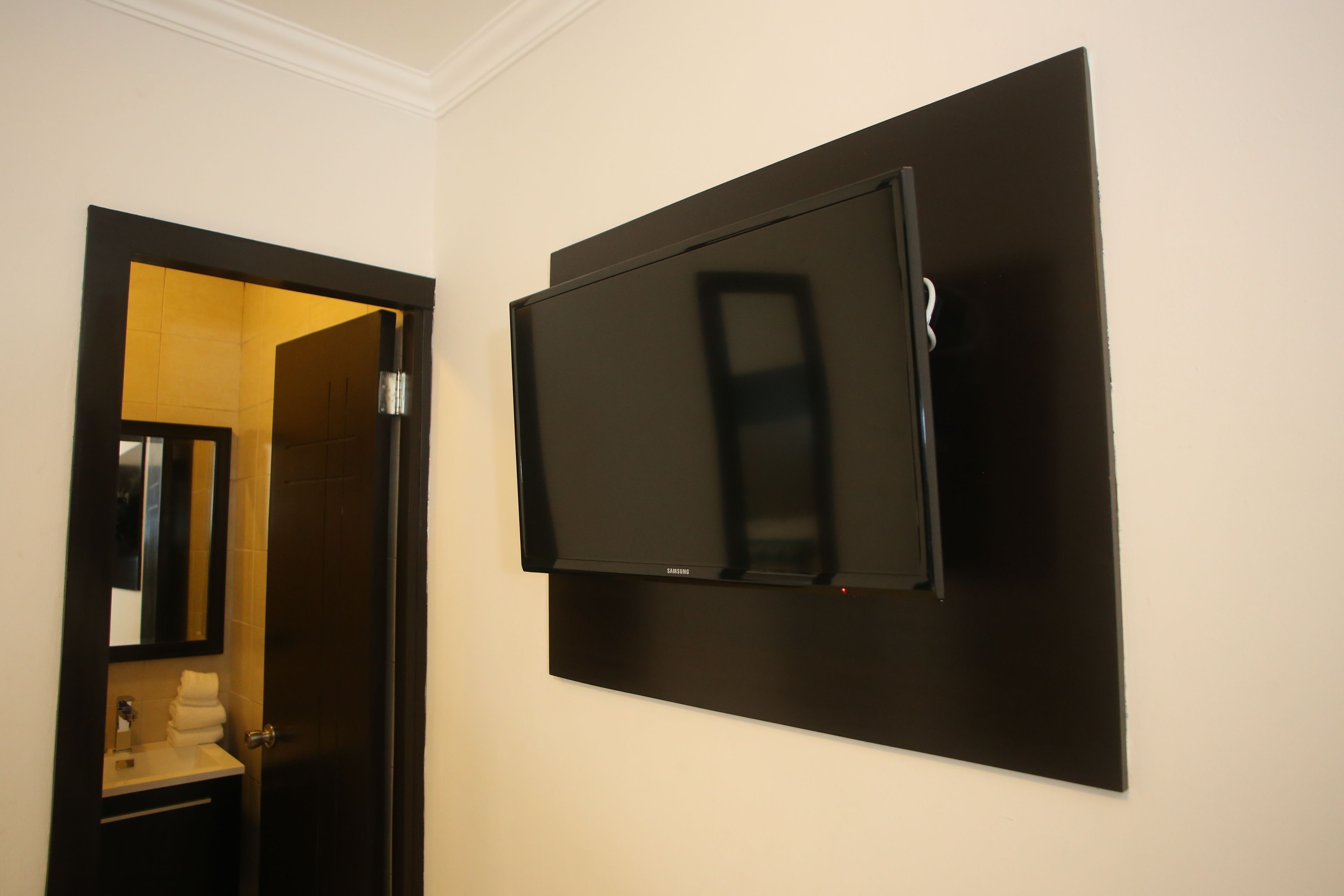 A room with a flat-screen TV mounted on the wall, a door leading to a bathroom, and a small sink with a mirror.