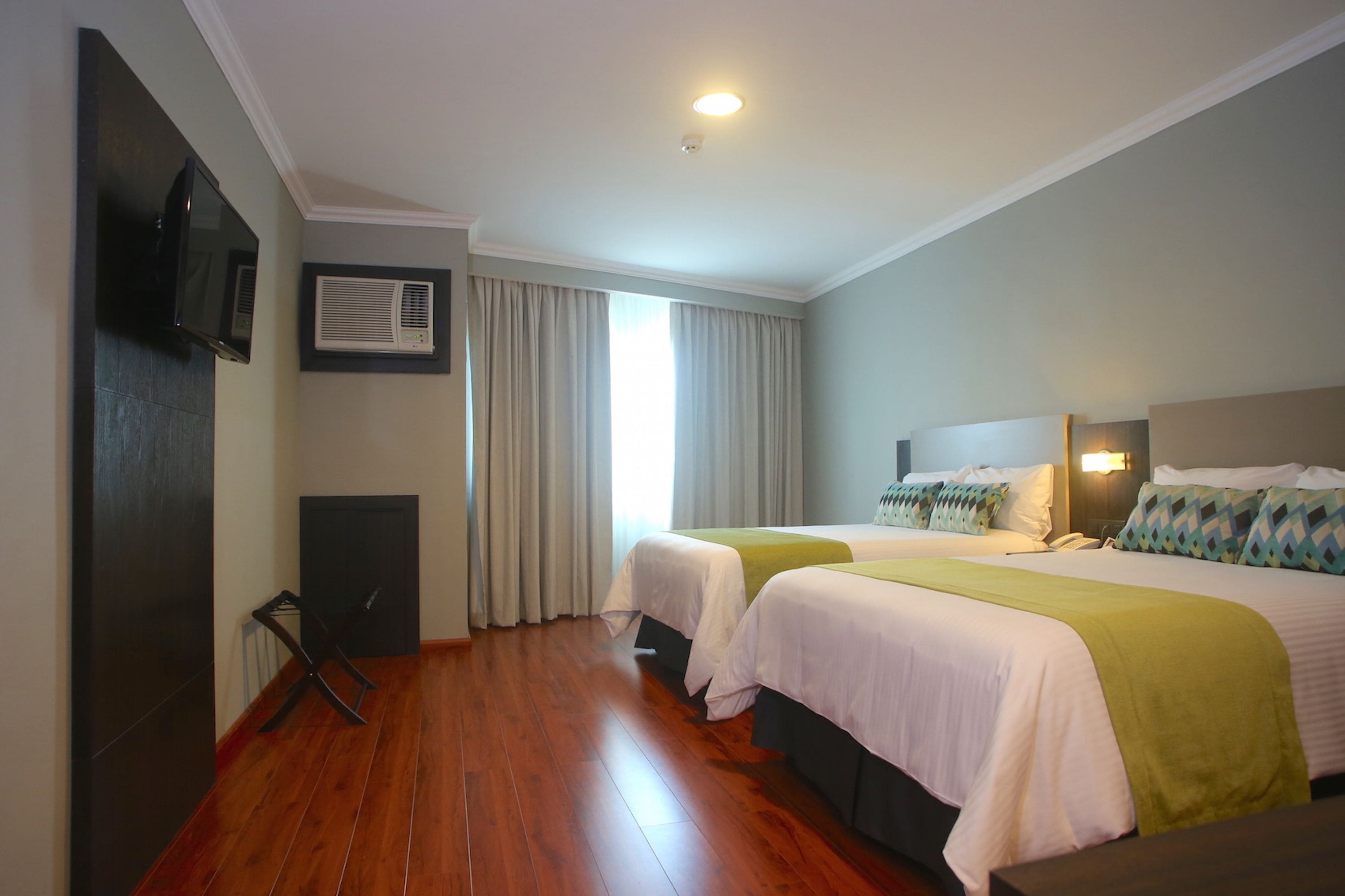 A hotel room with two beds, a flat-screen TV, an air conditioning unit, and a window with curtains.