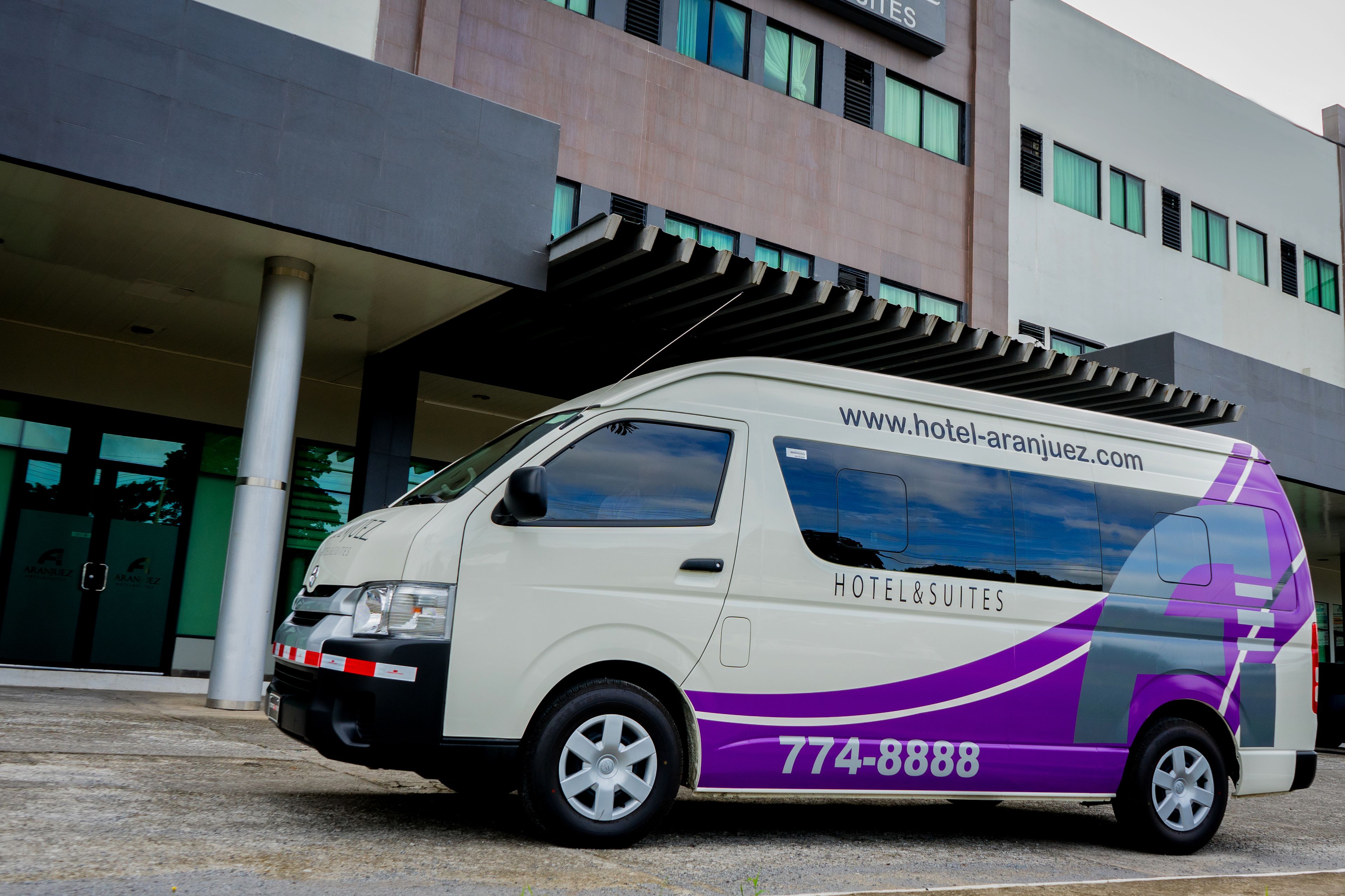 Uma carrinha branca e roxa estacionada em frente a um hotel, com o site www.hotel-aranjuez.com e o número 774-8888 visíveis na lateral.