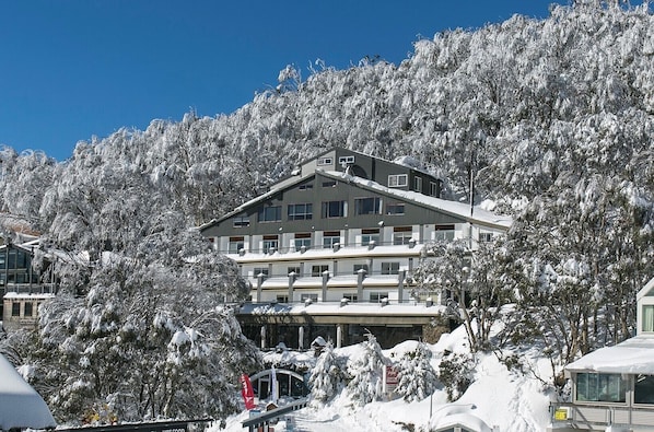 Un hotel de varios pisos con un diseño arquitectónico moderno, rodeado de árboles cubiertos de nieve y un cielo azul despejado.
