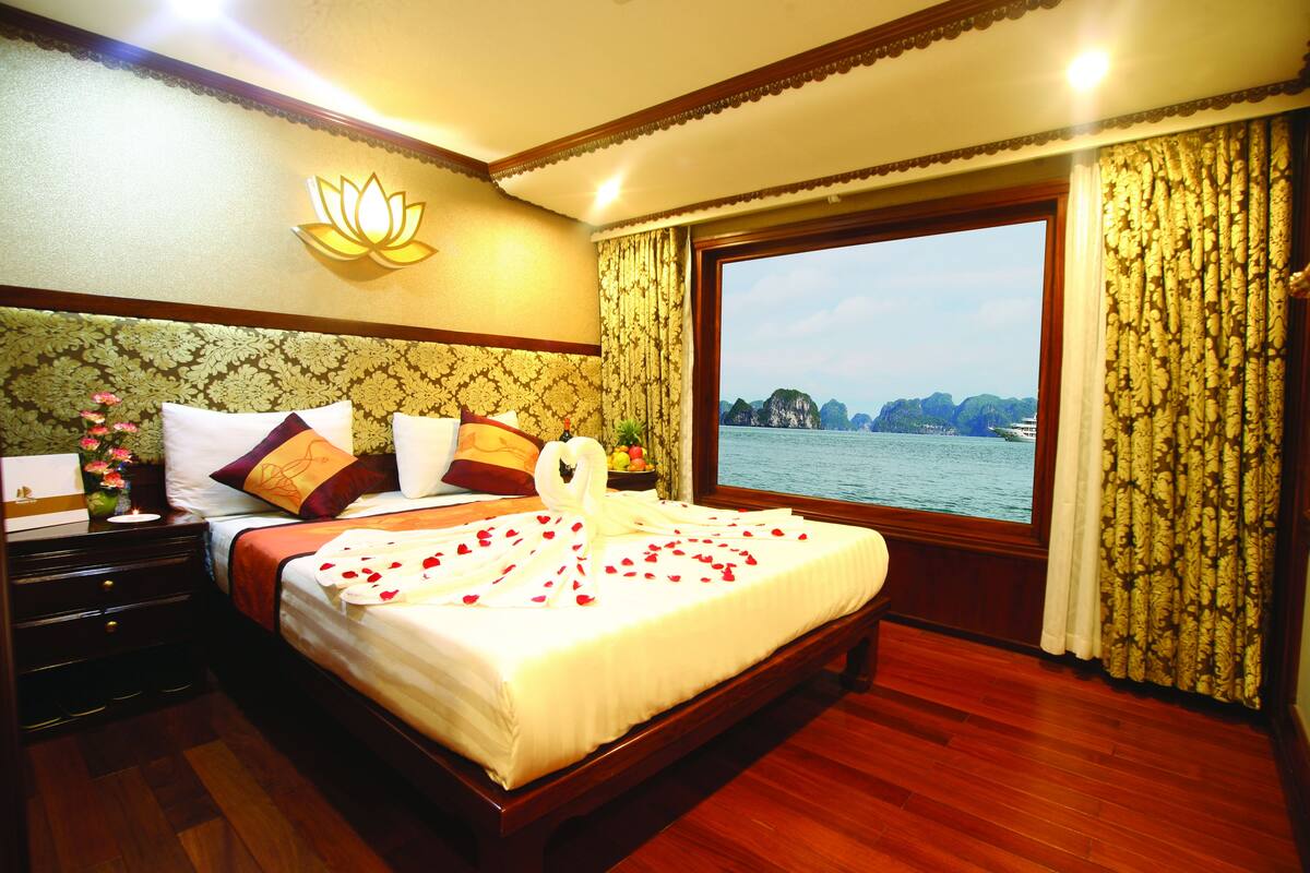 A hotel room with a large bed, a nightstand, a vase with flowers, and a window with a view of the sea and mountains.