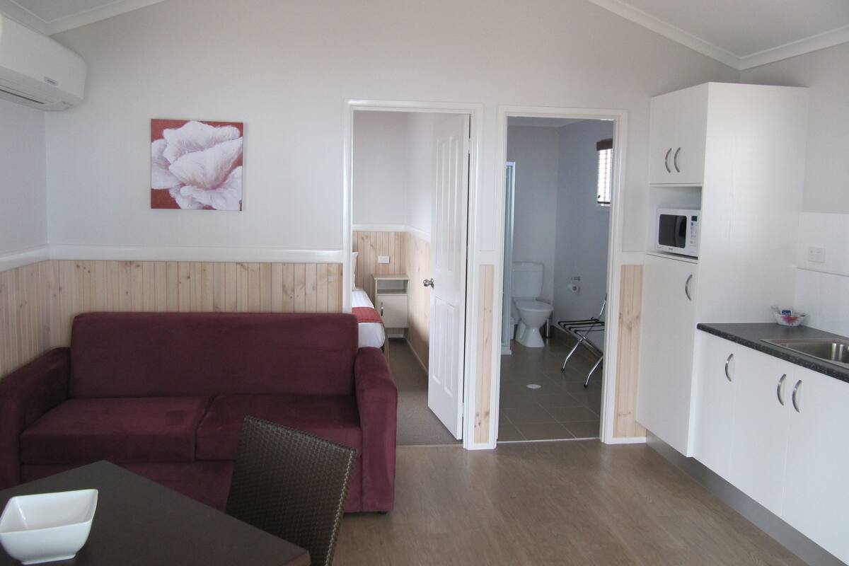 Un espacio habitable compacto con sofá, mesa de comedor, área de cocina con armarios y microondas, y un baño visible a través de un puerta abierta.