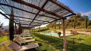 Un área de estar al aire libre con techo, sillones reclinables, piscina y jardín ajardinado.