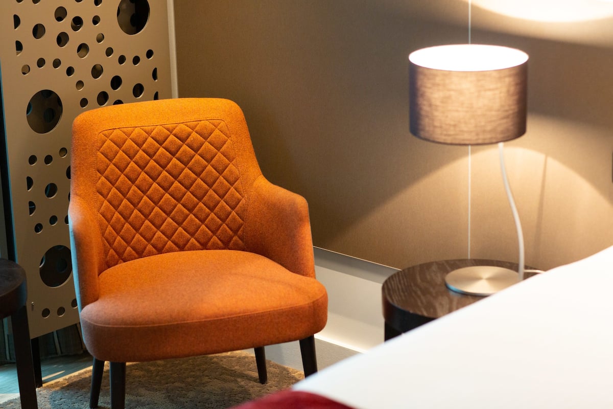 A modern room with a quilted orange armchair, a perforated wall, and a lamp with a cylindrical shade.