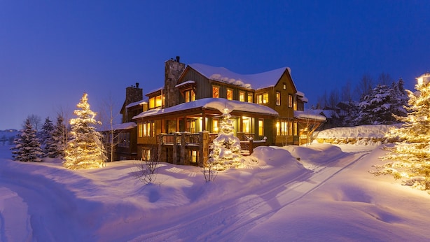 Un grand chalet à deux étages, dont le toit est recouvert de neige et qui possède de nombreuses fenêtres, entouré de neige et d’arbres.