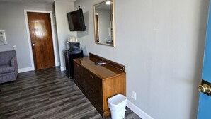 A room with a wooden dresser, a mirror, a television mounted on the wall, a gray sofa, and a trash can.