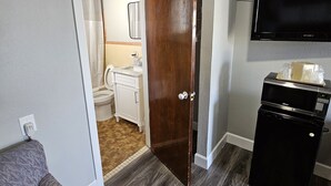 A compact living space with a grey sofa, a wooden door leading to a bathroom, a black refrigerator, and a mounted TV.