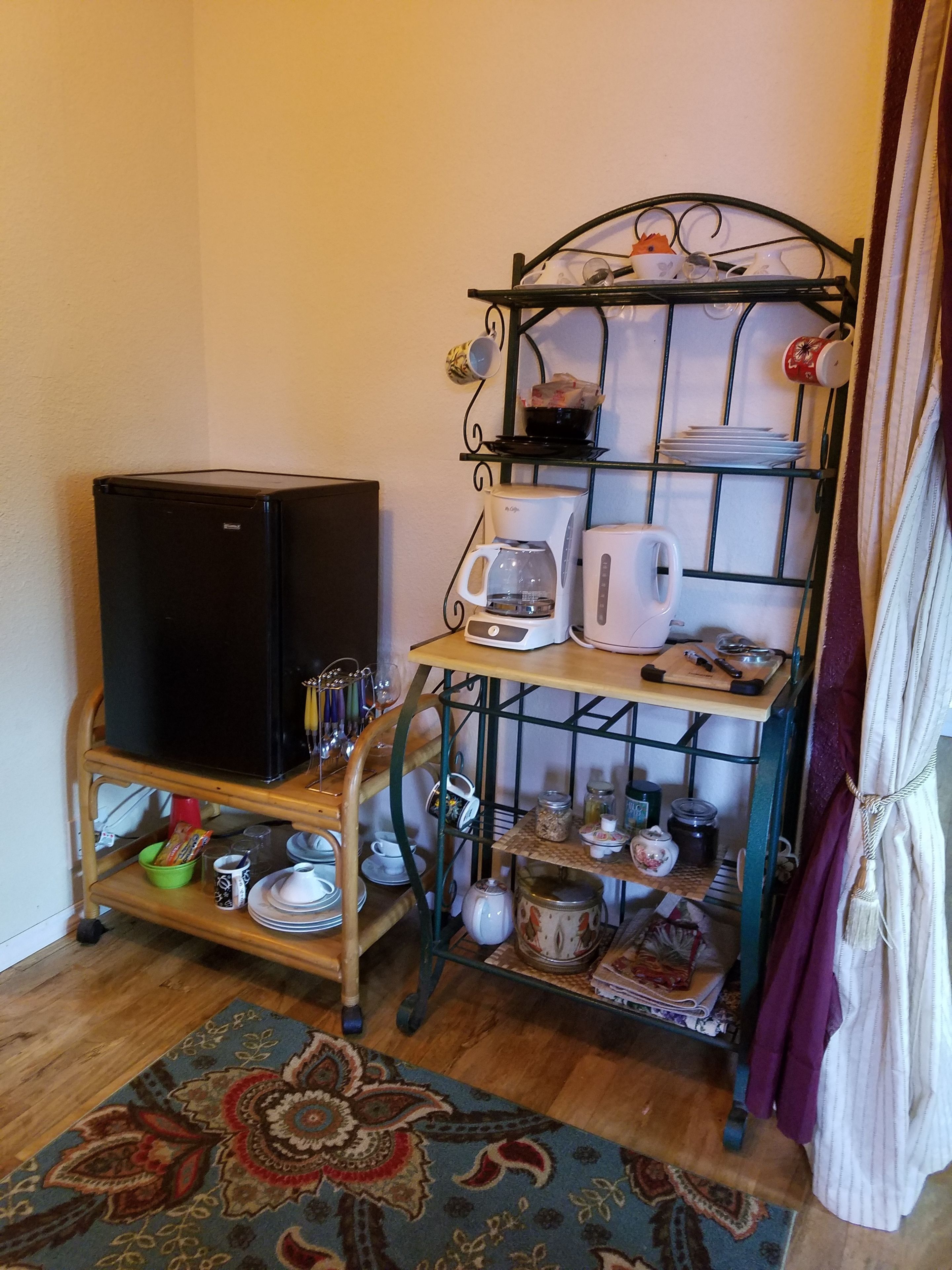 Una cocina compacta con refrigerador, cafetera, tostadora y una estantería metálica con vajilla y utensilios.