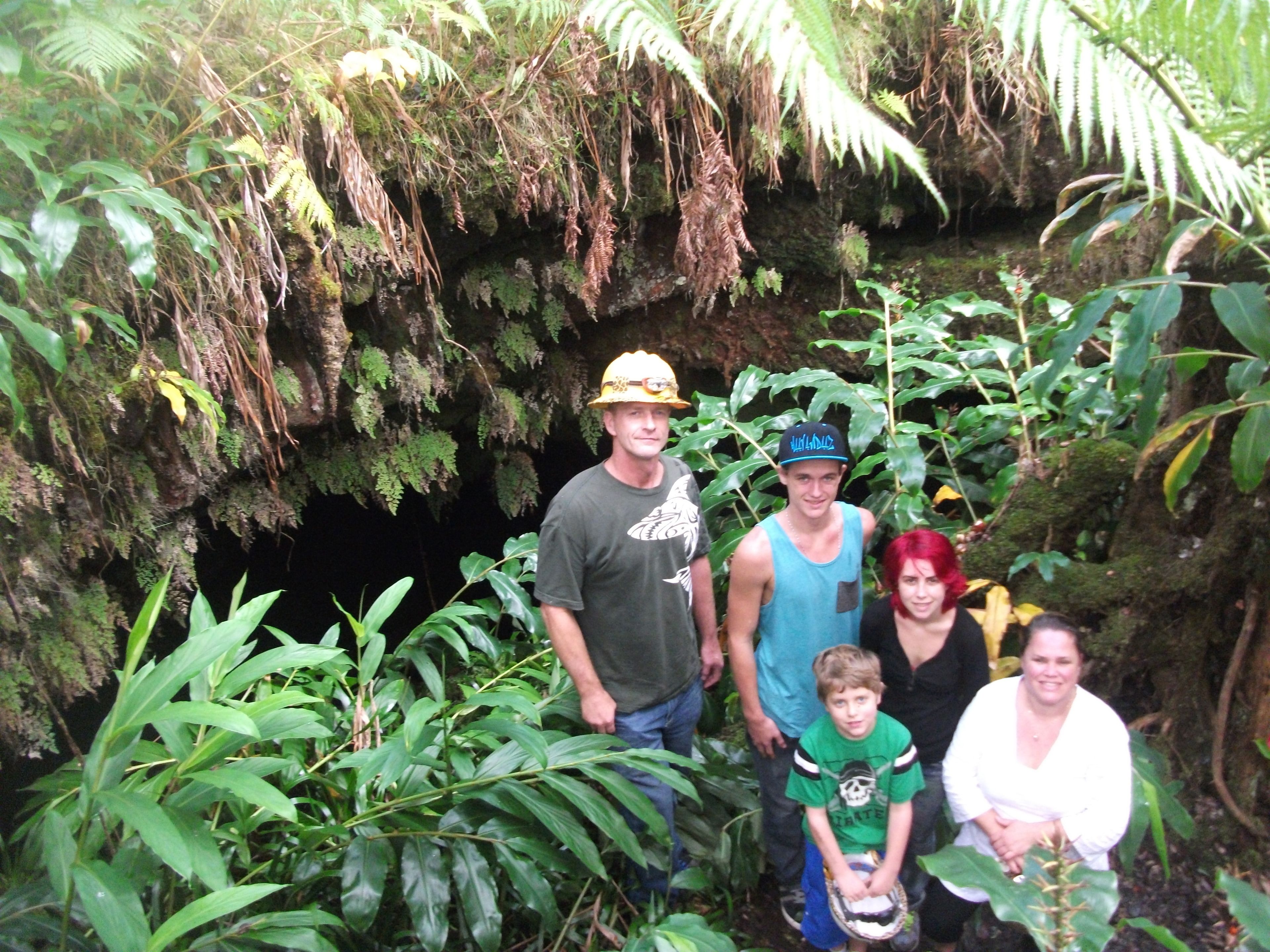 A group of people standing in a lush, green forested area with dense foliage and a rocky cave entrance.