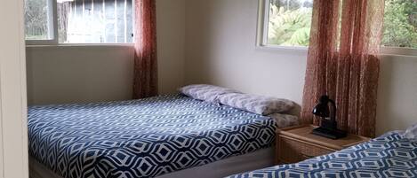 A room with two beds, a window with curtains, a small table with a lamp, and a view of a building outside.