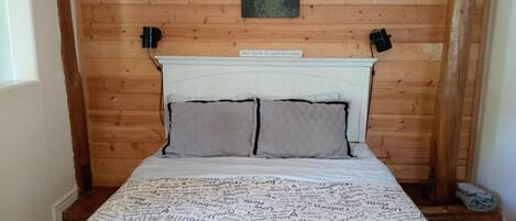 Une chambre à coucher aux murs lambrissés, avec une tête de lit blanche, deux oreillers et une couvre-lit ornée de motifs textuels.