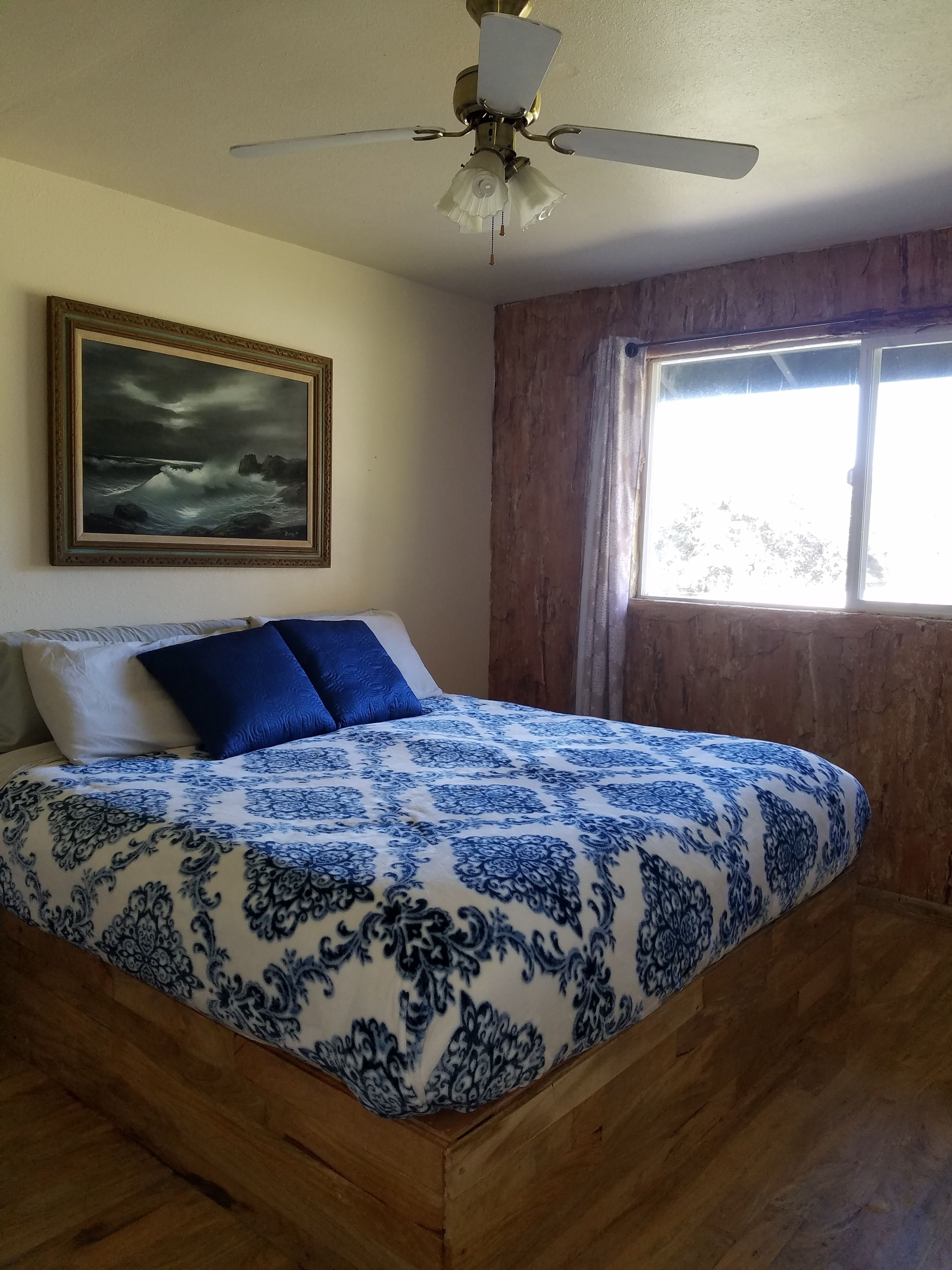 Une chambre à coucher avec un lit, un ventilateur de plafond, une fenêtre et un tableau au mur.