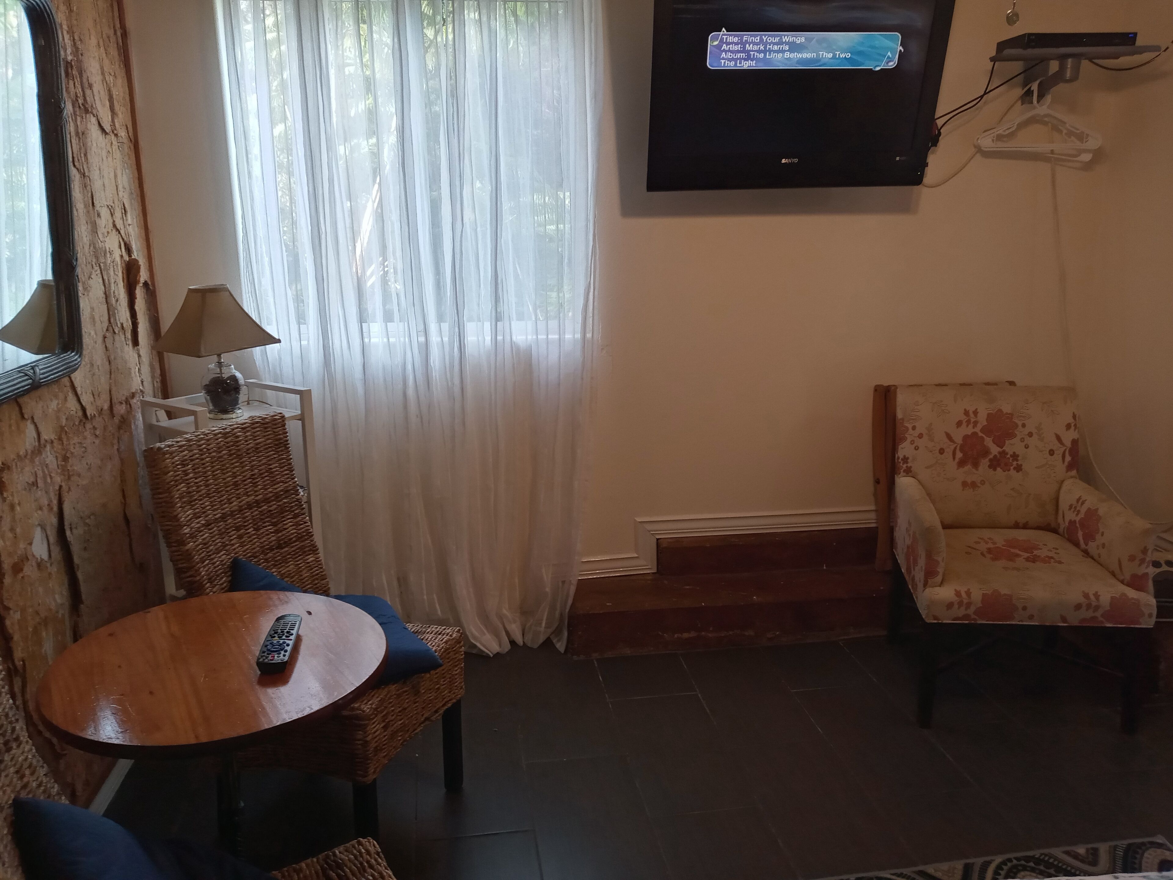 Una sala de estar con una silla de mimbre, una mesa redonda de madera, un control remoto y un televisor instalado en la pared.