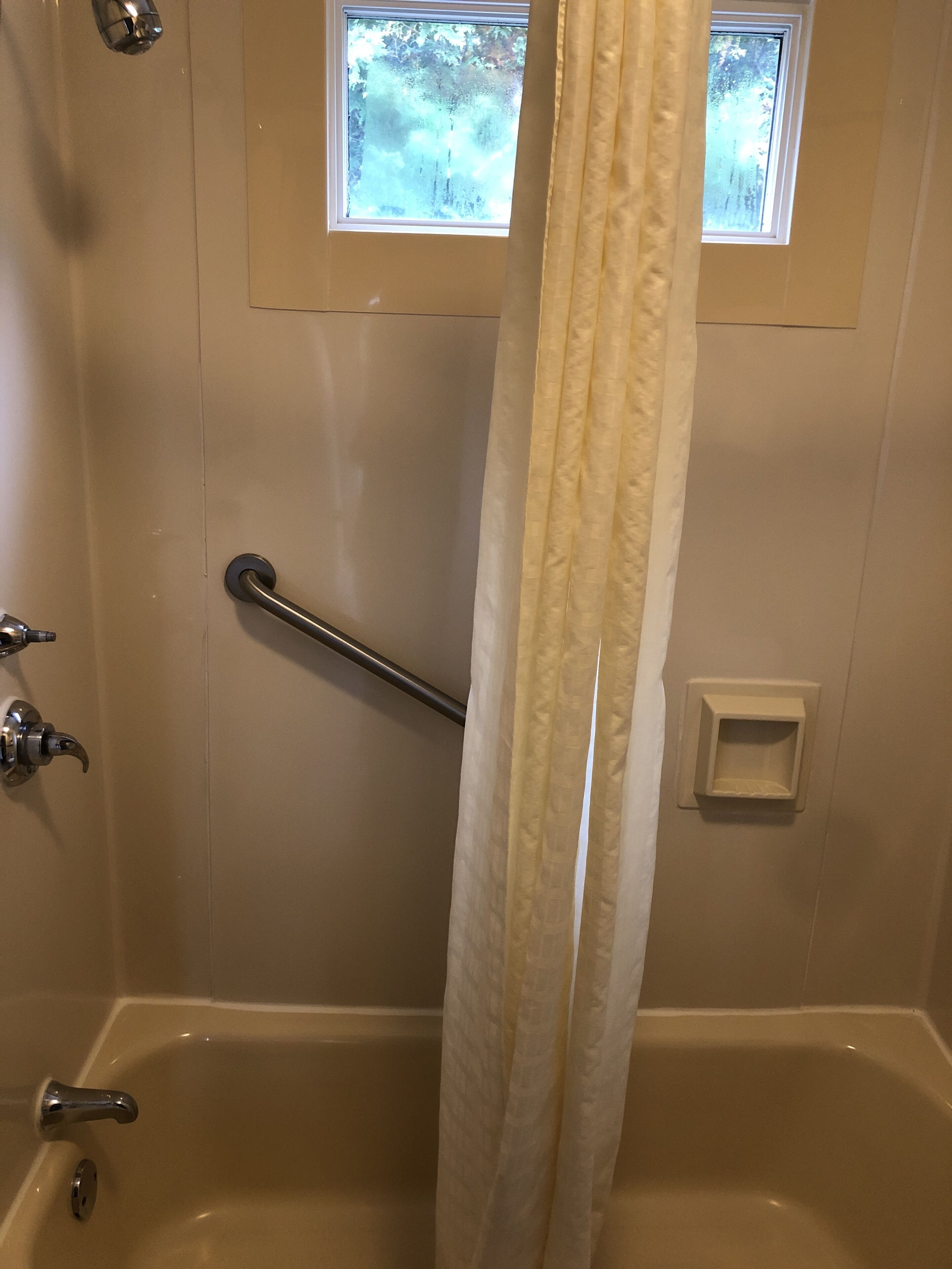 A bathroom with a bathtub, a shower curtain, a handrail, a window, and a small shelf.