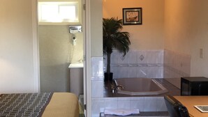 A hotel room with a bed, a bathtub, a sink, a mirror, a plant, and a framed picture on the wall. - Wilbraham Inn (Wilbraham)