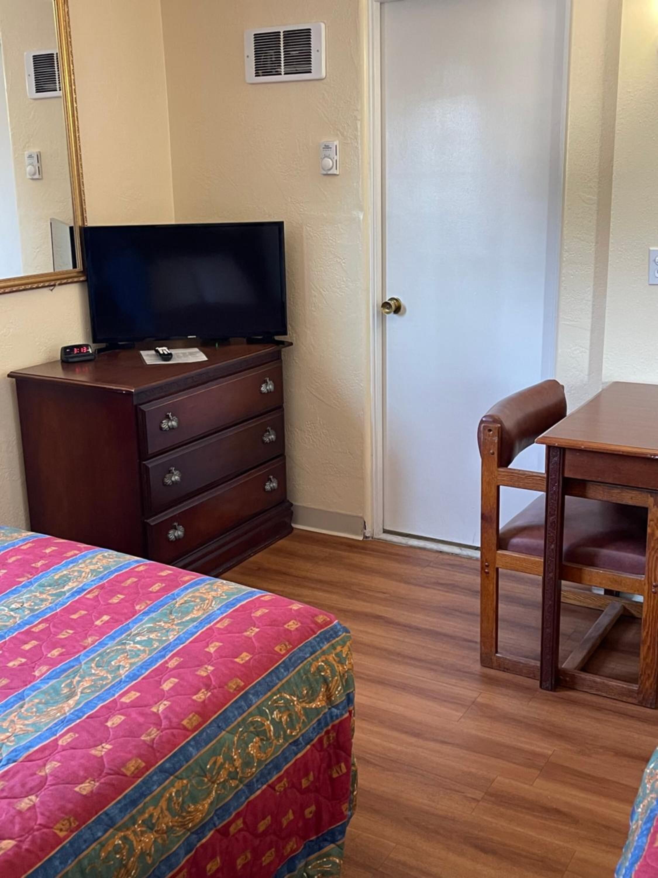 Quarto de hotel com cômoda de madeira, televisão, cama com colcha colorida, cadeira de madeira e uma porta.