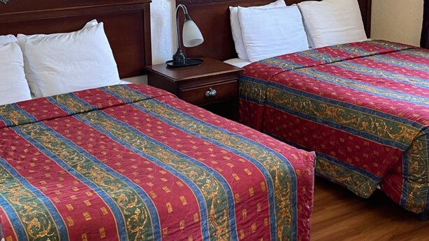 A hotel room with two beds, each with a red and gold patterned bedspread, a wooden headboard, and a nightstand with a lamp. A framed picture of a lighthouse is on the wall. - Villa West Motel (Florence)