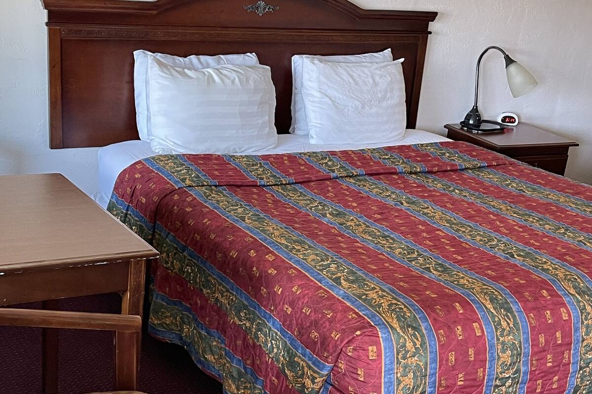 Quarto de hotel com cabeceira de madeira, cama com almofadas brancas, colcha com padrão em vermelho e verde, mesa de cabeceira de madeira com abajur e cadeira de madeira com enchimento estampado.