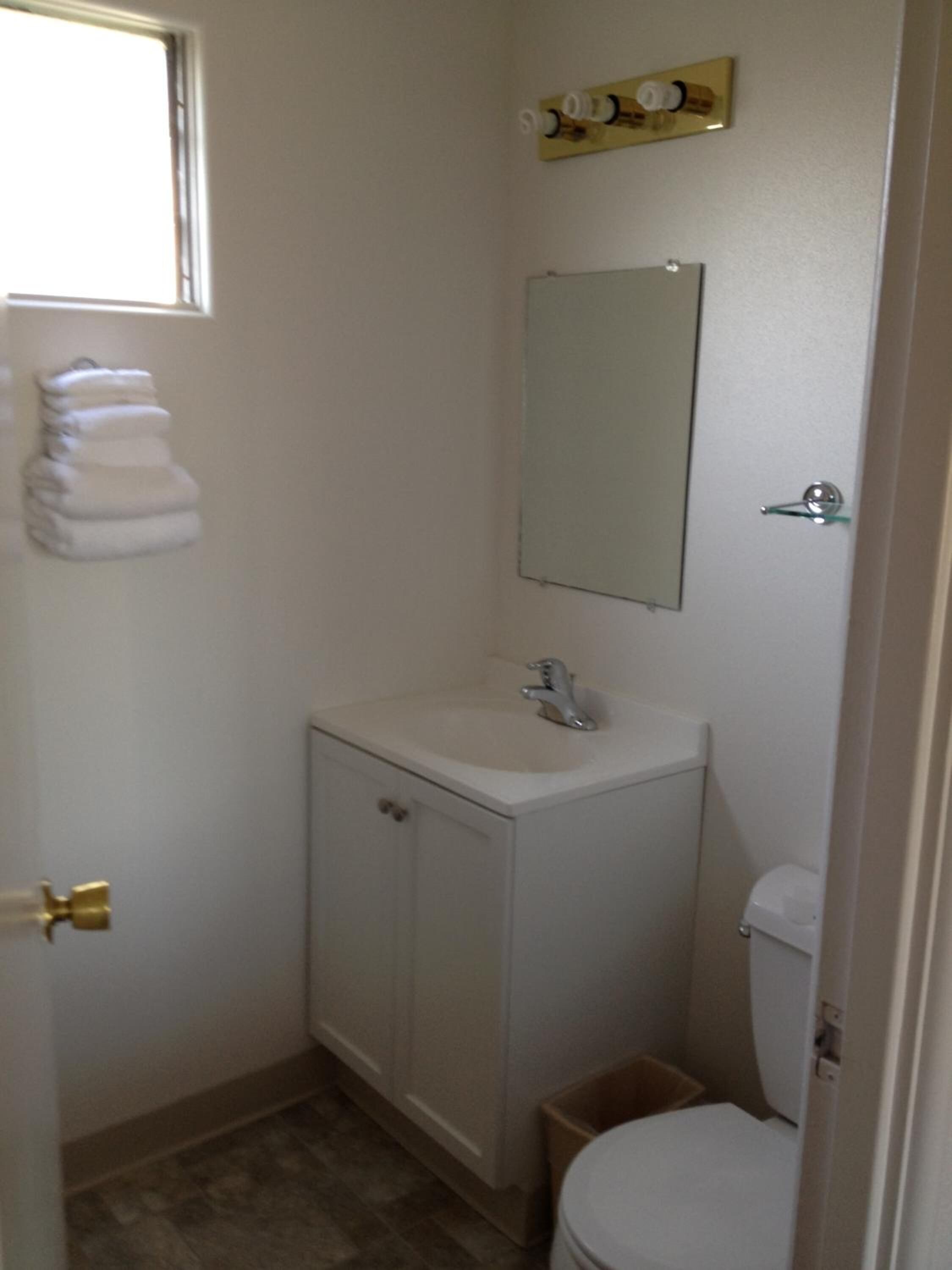 Un baño pequeño y funcional con un lavabo blanco, un espejo y un inodoro. Hay una ventana sobre el lavabo y un perchero en la pared.