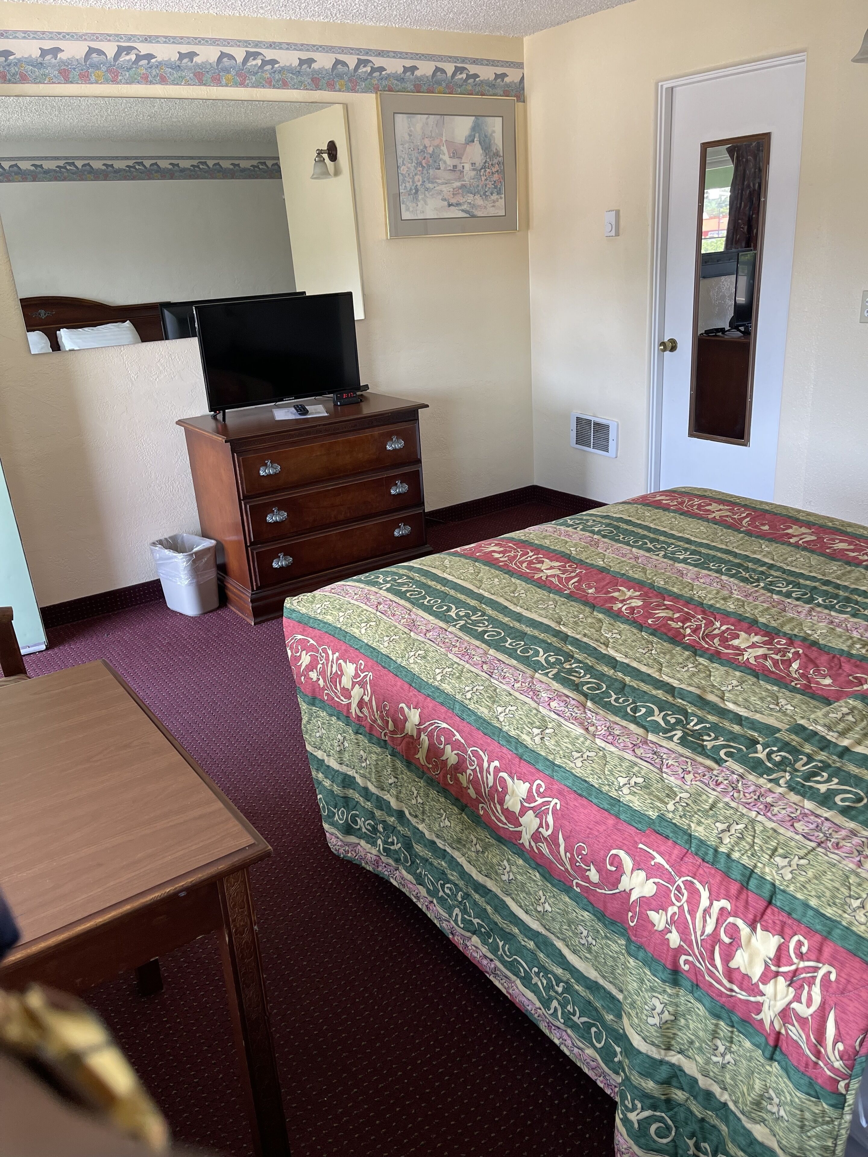 A hotel room with a bed, a wooden dresser, a television, and a door with a mirror.