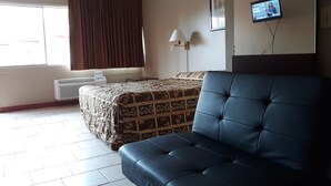 A hotel room with a bed, a blue armchair, a desk with a lamp, and a TV on the wall. - Longview Inn (Longview)