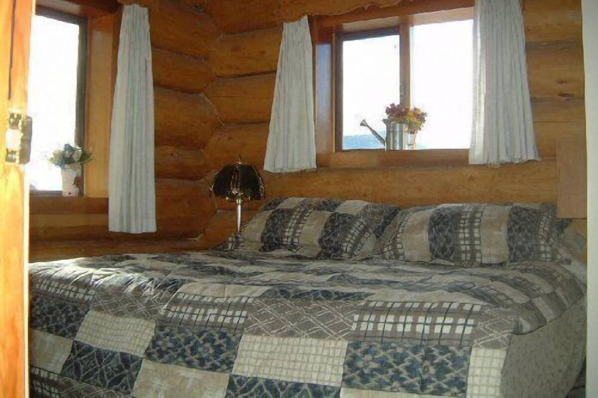 A log cabin bedroom with a quilted bedspread, two windows with curtains, and a lamp on the nightstand.