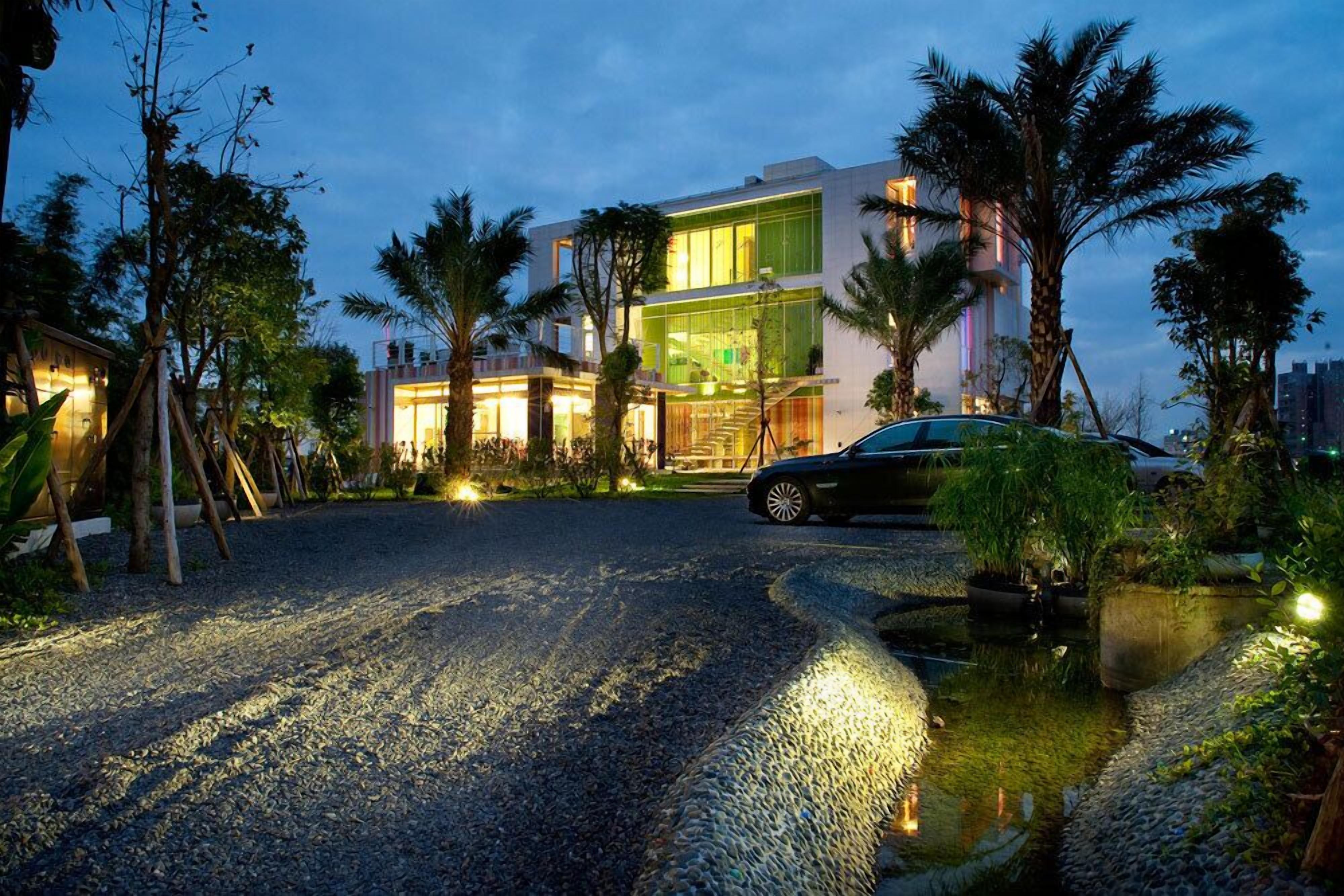 A modern building with a well-lit entrance, surrounded by palm trees and a gravel driveway leading to a car.