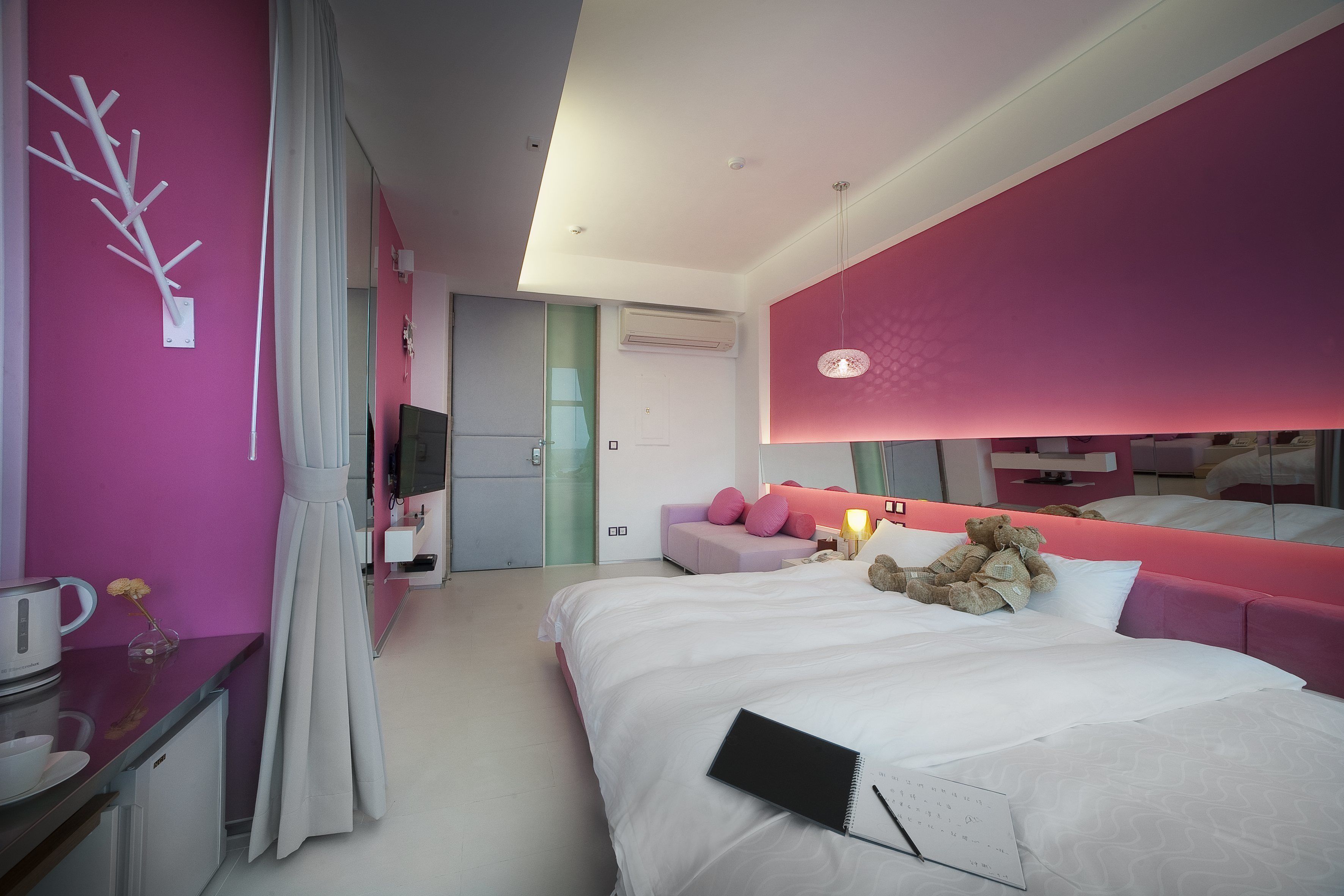 A modern hotel room with a large bed, a desk with a kettle, a mirror, and a teddy bear on the bed.