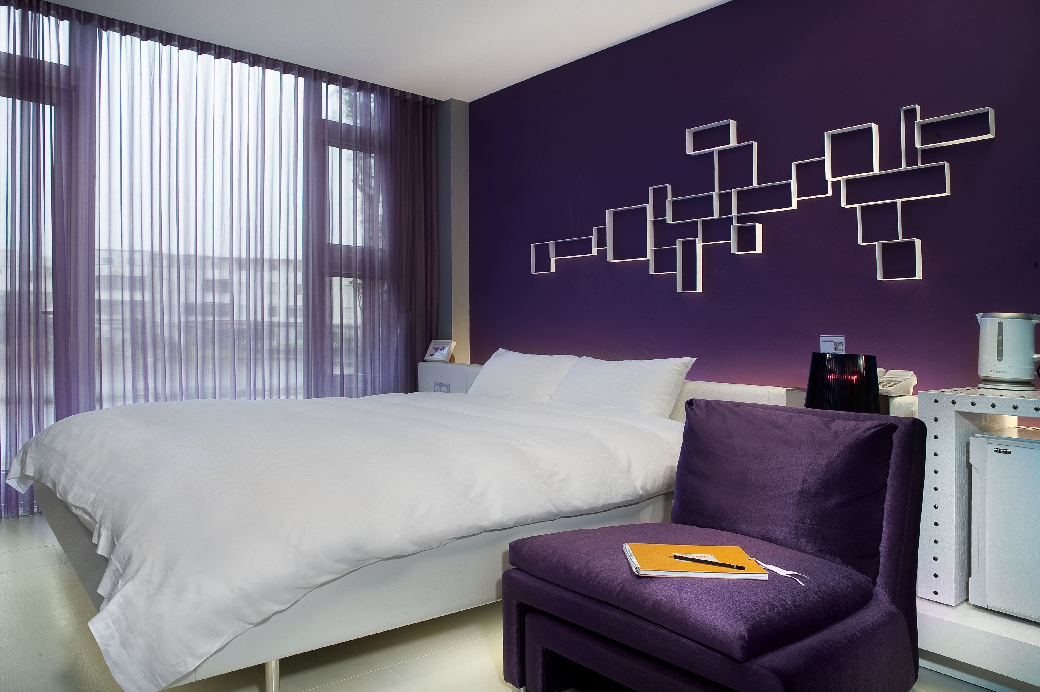 A hotel room with a bed, a purple armchair, a desk with a lamp, and a wall art piece.