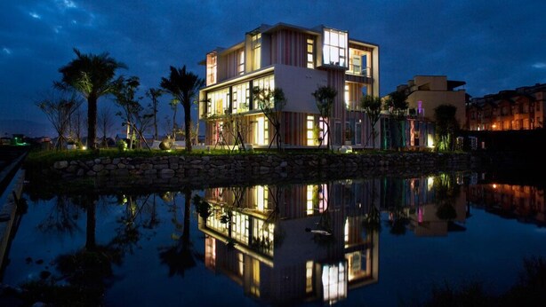 A modern multi-story hotel with a reflection in a body of water, featuring a stone pathway and palm trees. - La Palette (Luodong)