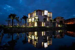 A modern multi-story hotel with a reflection in a body of water, featuring a stone pathway and palm trees.