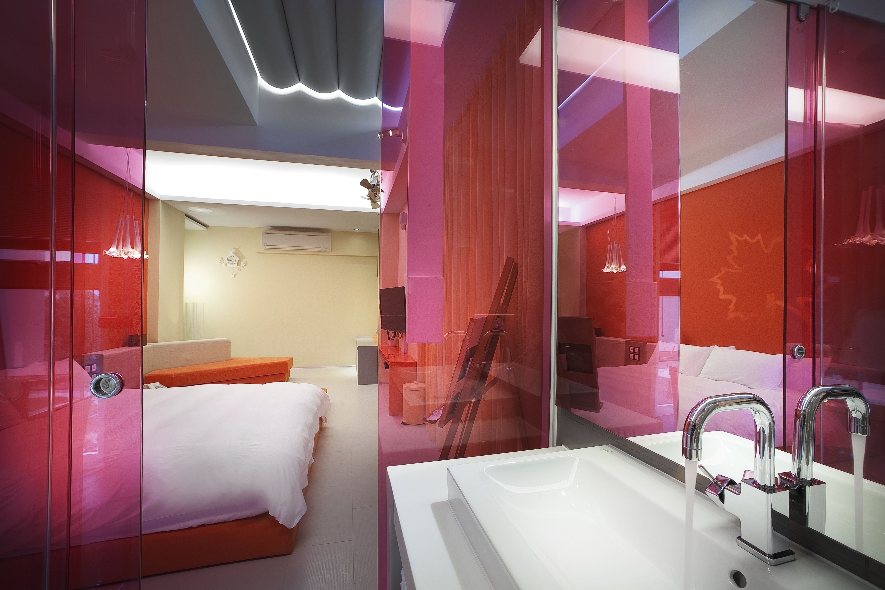 A modern hotel room with a glass partition, a white sink, and a bed with an orange headboard.