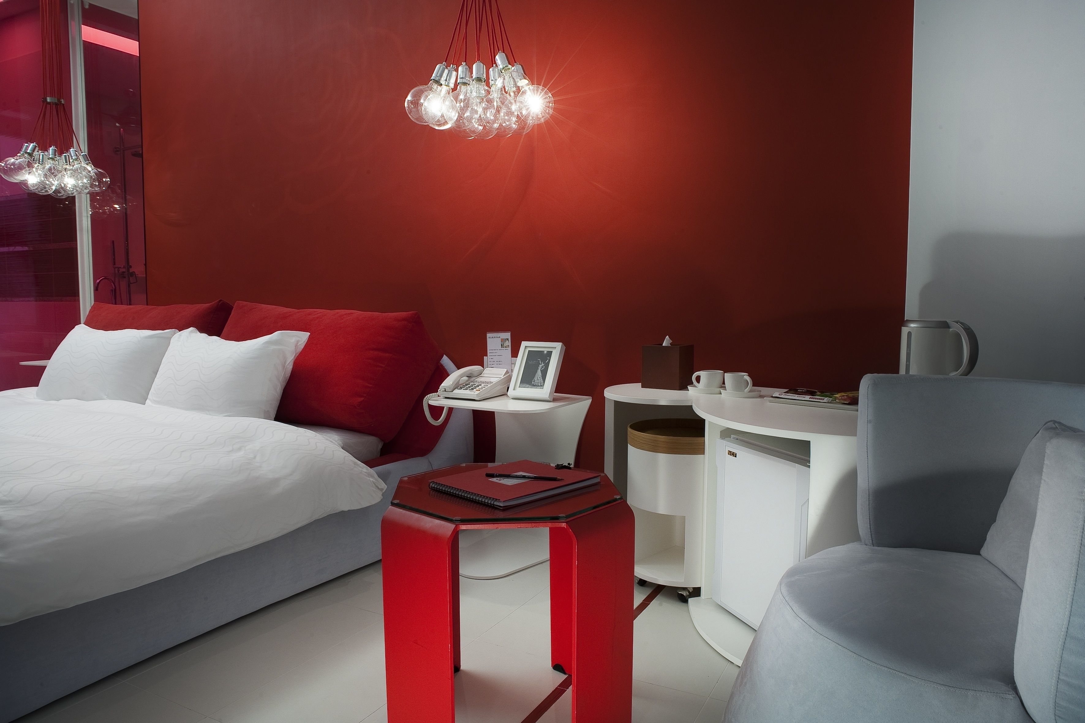 A modern hotel room with a bed, a desk, a chair, a red stool, and a chandelier.
