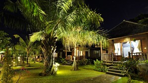 A well-lit tropical resort with a thatched-roof building, a paved pathway, and lush greenery. - Aman Gati Hotel Balangan (Jimbaran)