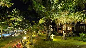A well-lit garden area with a pool, palm trees, and a wooden building. - Aman Gati Hotel Balangan (Jimbaran)