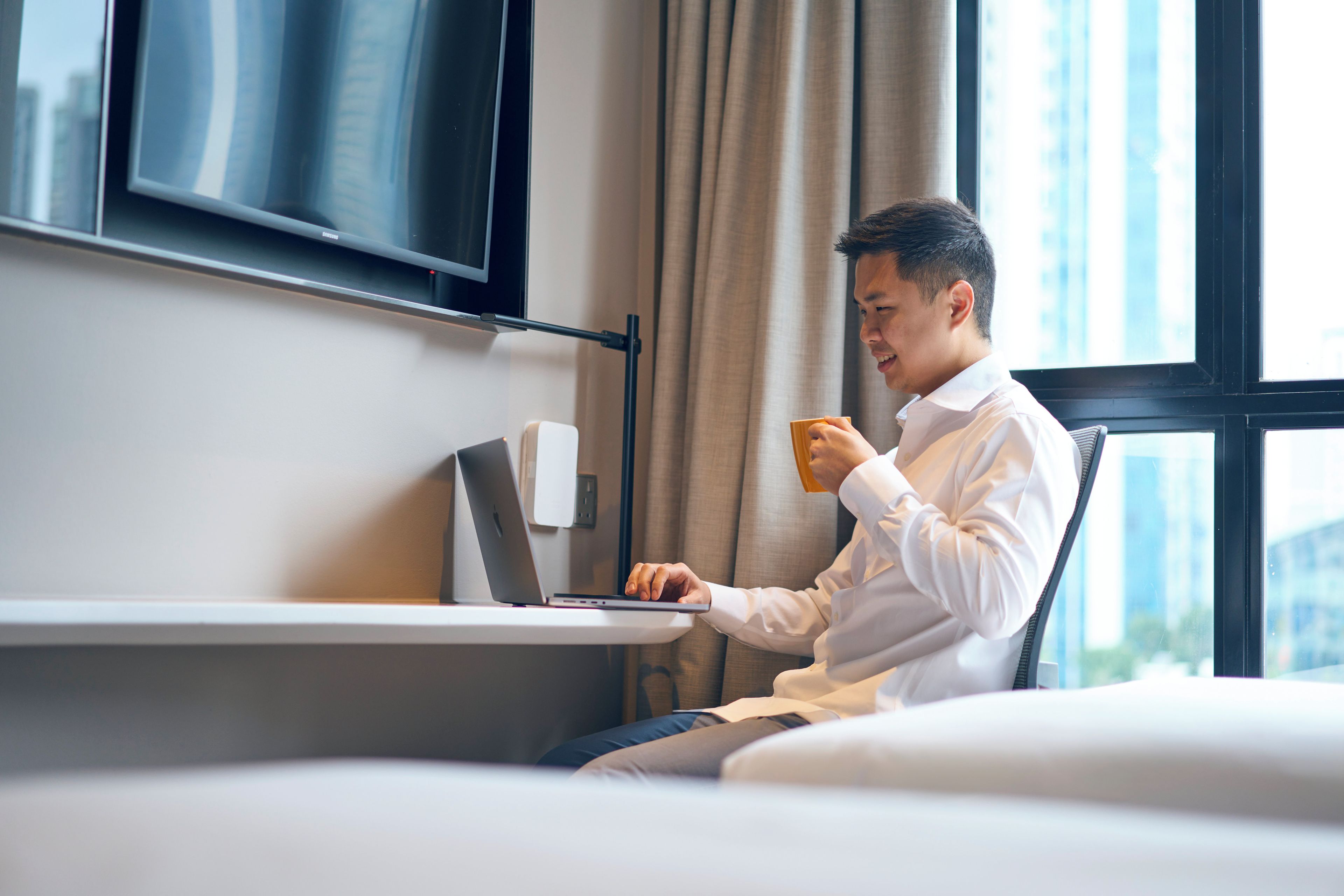 Una persona sentada en un escritorio con una computadora portátil, sosteniendo una taza, en una habitación con un ventanal grande y un televisor de pantalla plana montado en la pared.