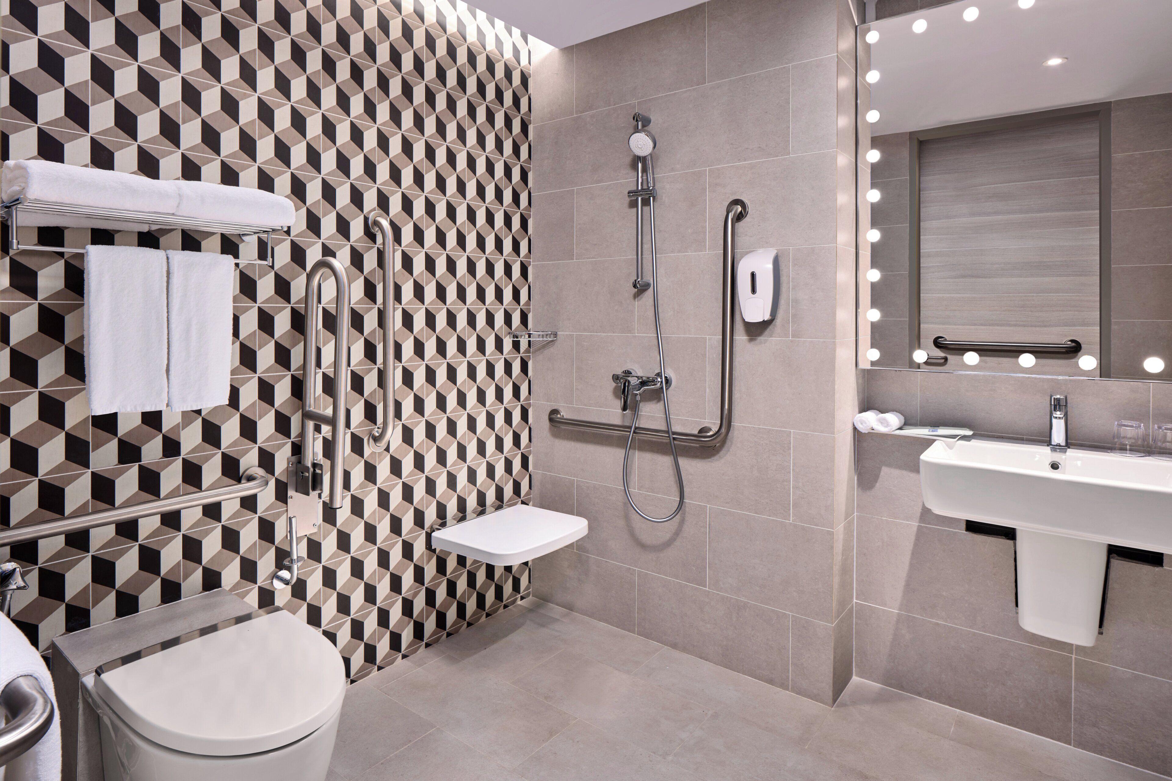 A modern bathroom with a geometric patterned wall, a toilet, a bidet, a shower with a handheld hose, and a vanity with a mirror and lights.
